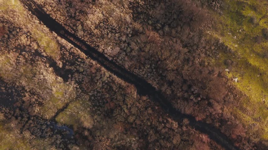 An aerial view shows a narrow stream flowing through a landscape of brown brush and green grass