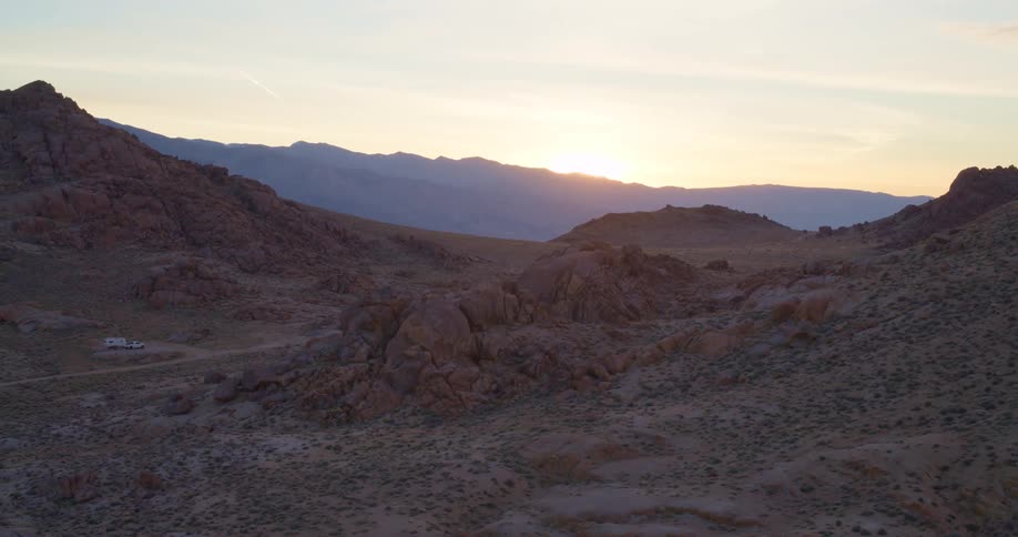 Golden sun sets behind distant mountains over a rocky desert landscape