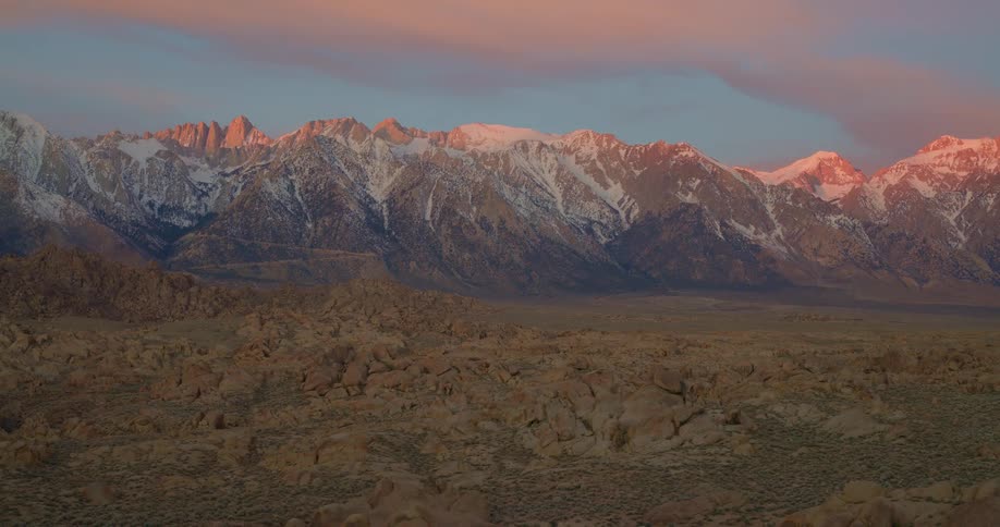 A view of a mountain range with peaks covered in snow shows a desert valley filled with rocks in the foreground during a sunset