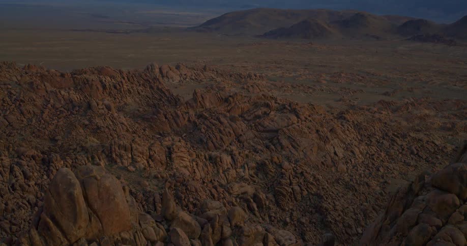 A vast rocky desert landscape viewed from an elevated perspective during the transition from sunset to dusk in the background a flat valley floor stretches toward a range of distant layered mountains under a fading pink and blue sky