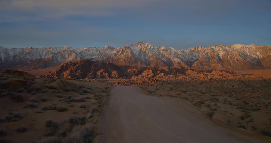 Aerial view moving along a dirt road shows a desert landscape leading toward a range of snowy mountains during a sunset