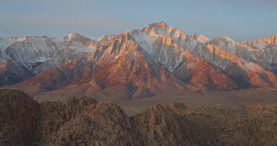 The golden light of sunrise illuminates the jagged snowdusted peaks of a massive mountain rangein the foreground rugged rock formations in deep shadow provide a sharp contrast to the