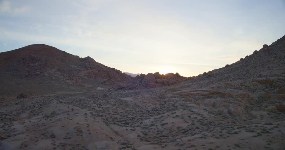 Plants and vegetation spread across the desert with a truck and camper parked at the foot of a mountain as the sun shines on the horizon