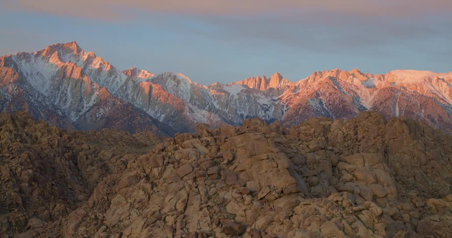 Golden sunlight illuminates the peaks of a snowcapped mountain range during a clear sunrisein the foreground a vast landscape of jagged tancolored rock formations sits in the early morning shadows