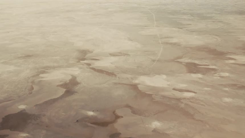 An aerial view of a vast flat desert or salt pan with intricate marbled patterns of light and dark sand a thin winding trail stretches across the parched earth disappearing into the hazy monochromatic horizon