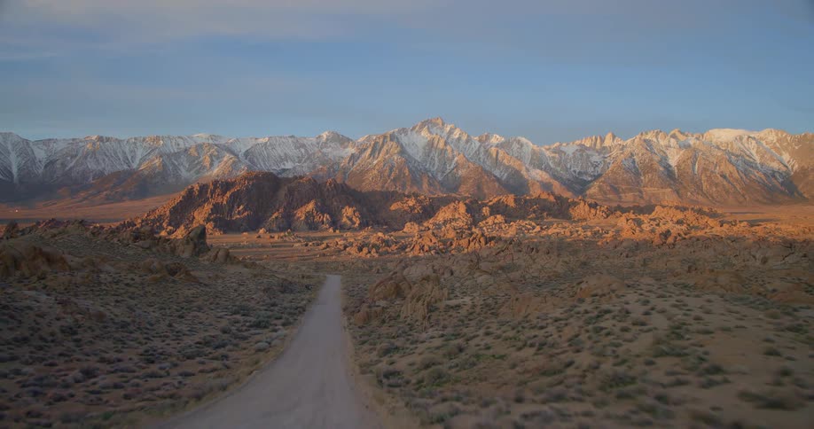 Dirt road crossing arid land toward sunlit snowy mountains