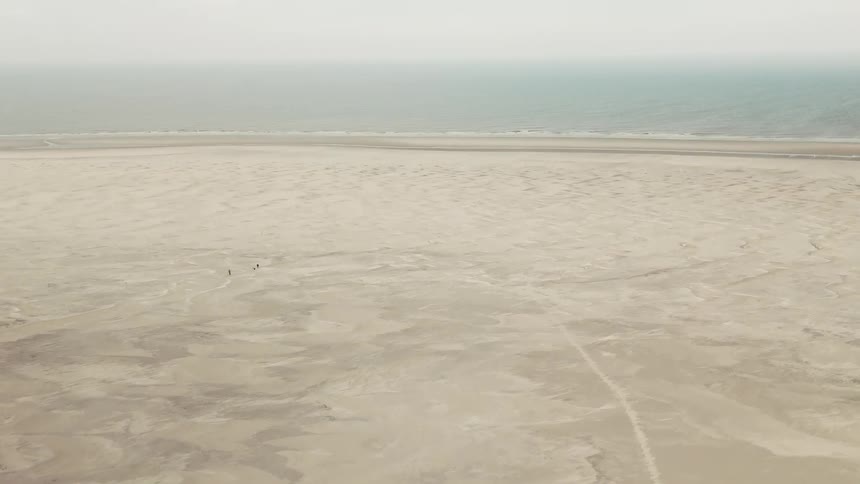 An aerial view shows a vast and flat sandy beach that stretches toward the ocean under a pale sky