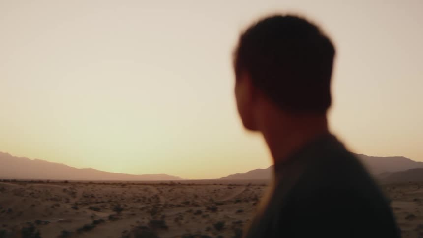 A view from behind a person shows them as they look out at a desert landscape under a warm sky at sunset