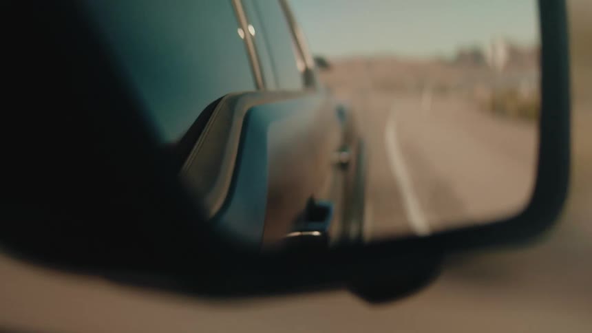 The video shows a closeup cinematic view of a desert road reflected in the side mirror of a moving vehicle the golden hour lighting creates a warm nostalgic atmosphere as the blurred landscape recedes into the distance