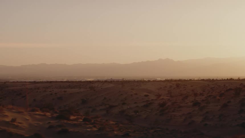A view of a desert landscape shows sand and small shrubs under a sky filled with a warm orange glow at sunset