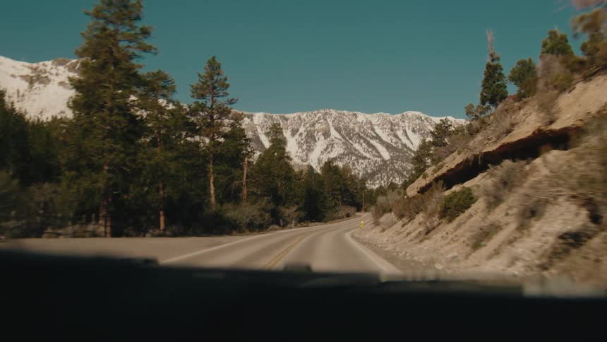 A view from a moving vehicle travels along a winding asphalt road through a pine forest toward large mountains covered in snow under a clear blue sky on a bright day