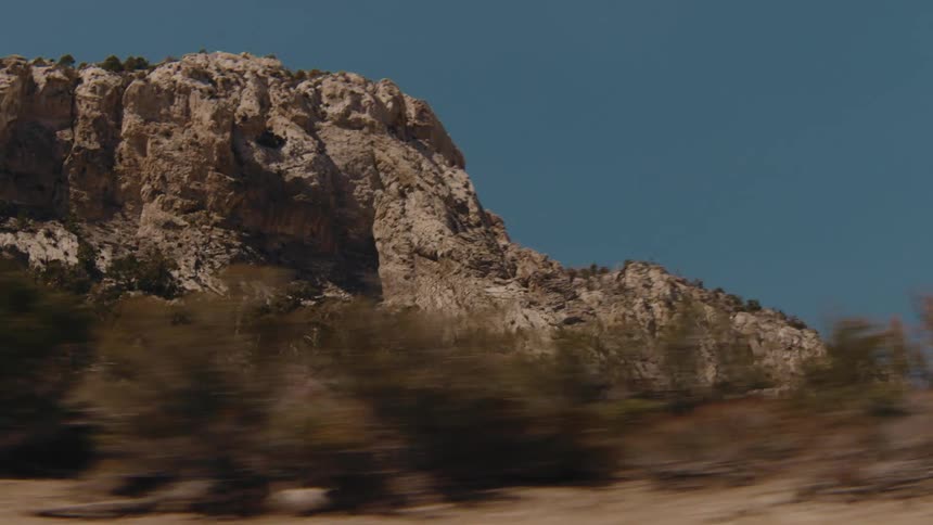 A view from a moving vehicle shows a large rocky mountain under a clear blue sky with dry brush and small trees passing quickly in the foreground during a sunny day