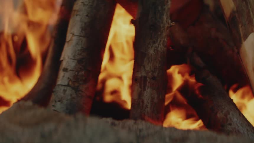 Bright orange and yellow flames dance between several thick pieces of wood arranged in a campfirethe fire burns steadily on a sandy surface with the glowing embers and heat creating a soft blur around the logs