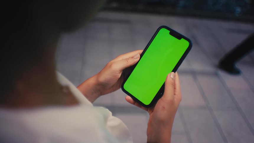A person stands outdoors in the evening and holds a smartphone with a green screen in a close view while people walk past them on a paved walkway in the background