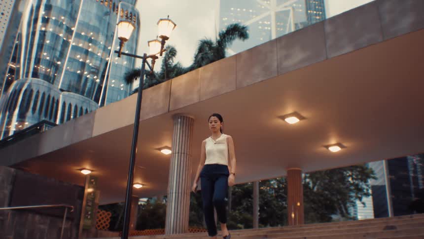 A woman wearing a white sleeveless top and dark pants walks down a set of stone stairs in a city plaza at dusk with illuminated modern buildings and palm trees in the background