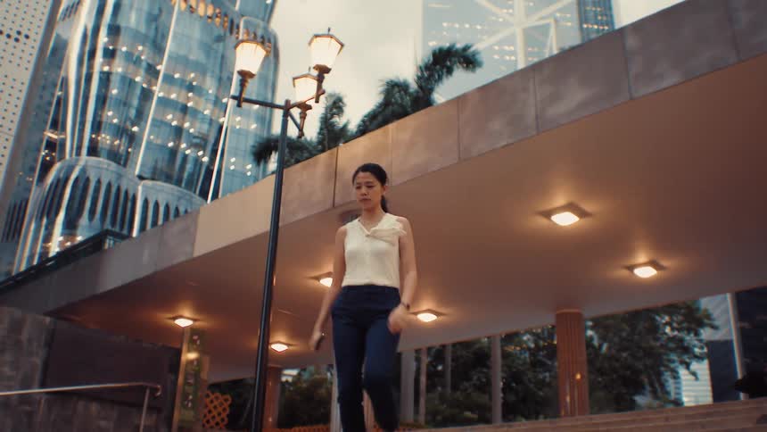 A woman wearing a white sleeveless top and dark pants walks down a set of stone stairs in a city plaza at dusk with illuminated modern buildings and palm trees in the background
