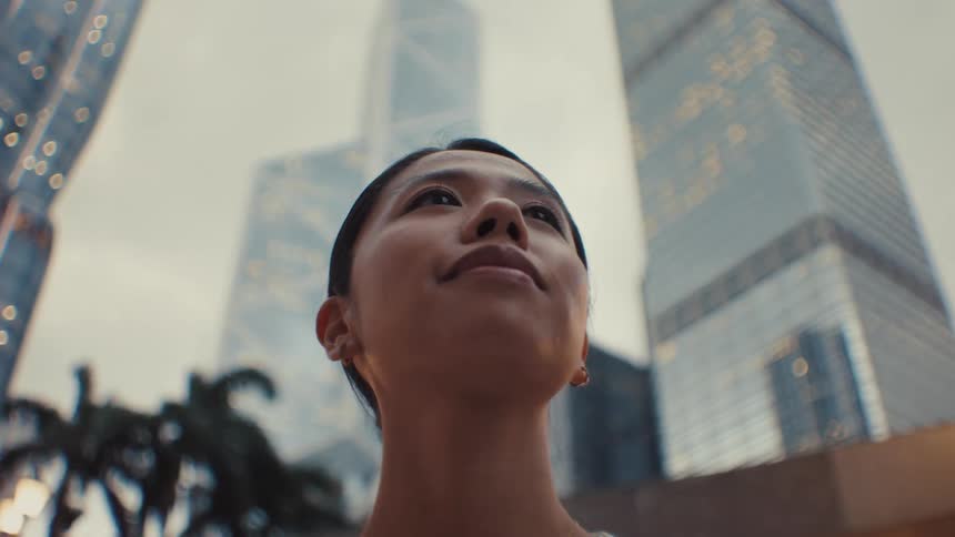 A lowangle view of a woman gazing upward against a backdrop of illuminated skyscrapers at dusk in a modern city