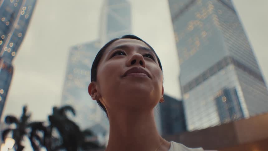 A lowangle view of a woman gazing upward against a backdrop of illuminated skyscrapers at dusk in a modern city