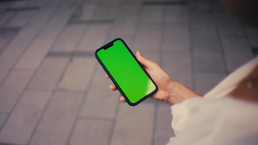 A woman wearing a white shirt stands on a paved city walkway while holding a smartphone with a green screen in a close view