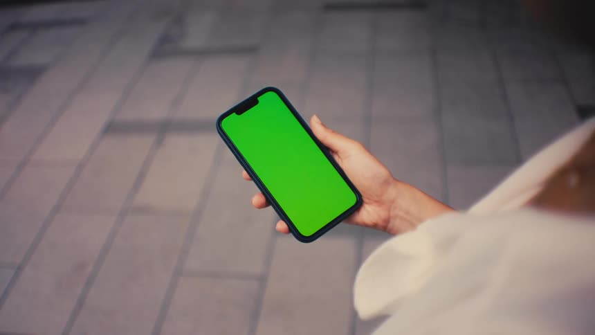 A woman wearing a white shirt stands on a paved city walkway while holding a smartphone with a green screen in a close view