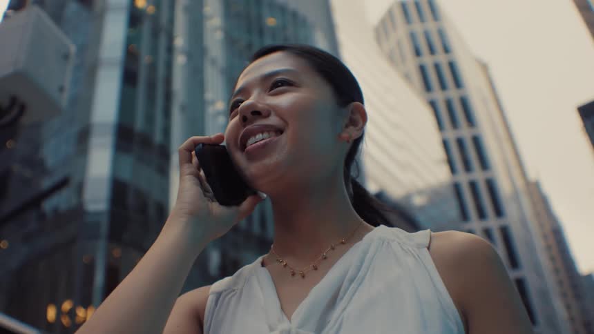 A woman in a white sleeveless blouse talks confidently through a canyon of tall glass skyscrapers in a modern city