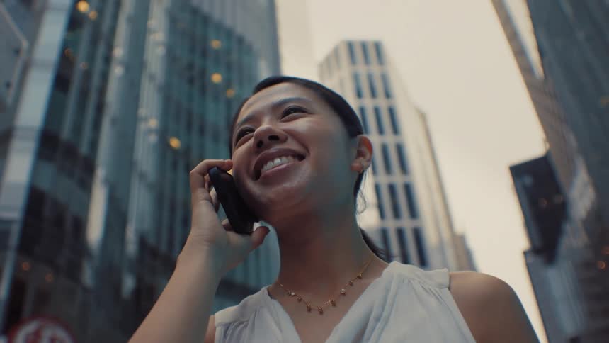 A woman in a white sleeveless blouse talks confidently through a canyon of tall glass skyscrapers in a modern city