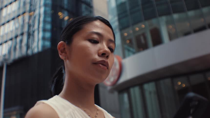 A woman wearing a white sleeveless top and a gold necklace walks through a city while talking on a mobile phone and looking at the modern glass buildings surrounding her during the day