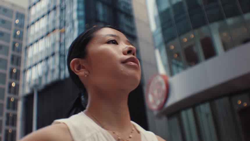 A woman wearing a white sleeveless top and a gold necklace walks through a city while talking on a mobile phone and looking at the modern glass buildings surrounding her during the day