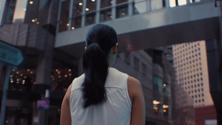 The camera follows a woman with dark hair tied in a ponytail as she walks through a dense urban environment highrise buildings and an elevated pedestrian bridge frame the scene as she moves through the city streets