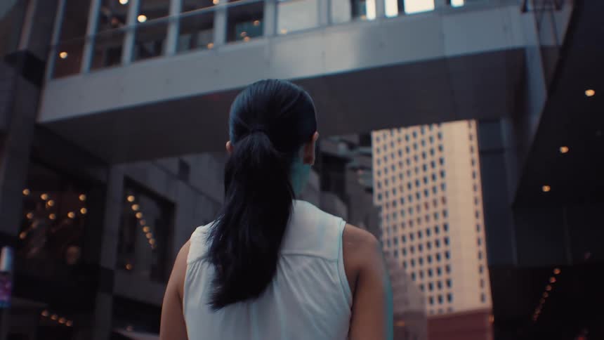 The camera follows a woman with dark hair tied in a ponytail as she walks through a dense urban environment highrise buildings and an elevated pedestrian bridge frame the scene as she moves through the city streets