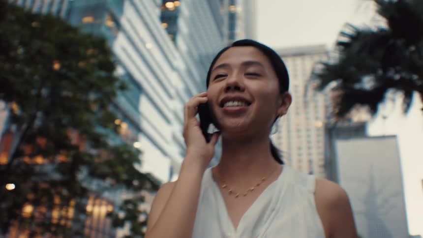 A woman wearing a white sleeveless top walks through a city with tall buildings while talking on a mobile phone and smiling in a low view