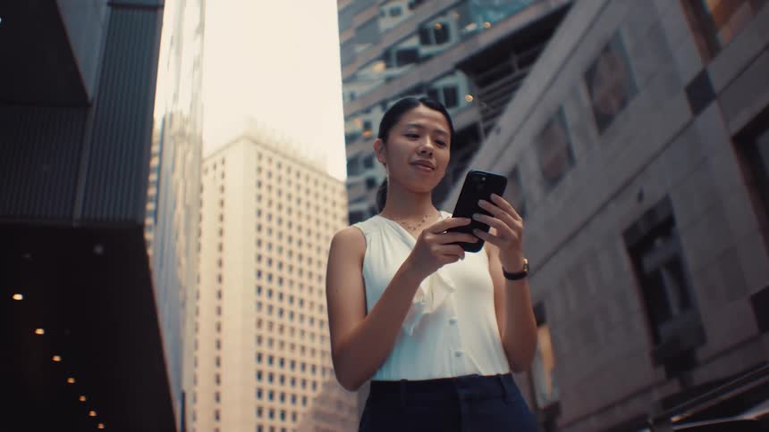 A woman wearing a white sleeveless top and dark trousers walks through a city with tall buildings stops to check her phone while smiling then puts the device in her pocket and continues walking