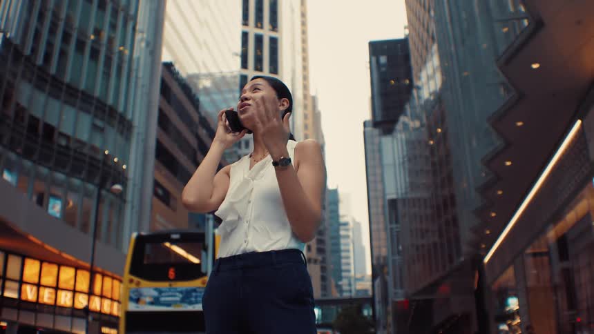 A woman in business attire stands on a city street between tall buildings smiling as she talks on her phone beside a passing doubledecker bus