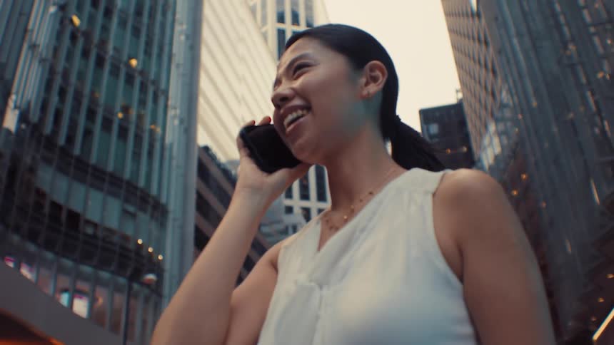 A smiling asian woman walks through a dense city district while talking on her smartphone