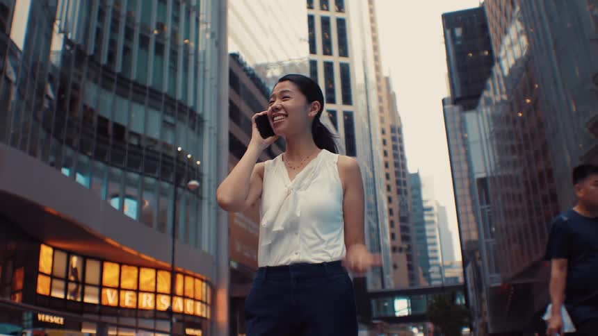 A woman wearing a white sleeveless top and dark pants walks through a city and smiles while talking on her phone with tall modern buildings and city traffic in the background