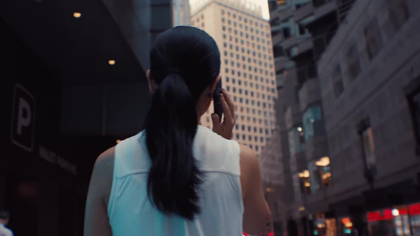 A woman with dark hair tied in a ponytail wearing a white sleeveless top walks through a city while answering a phone call in a view from behind