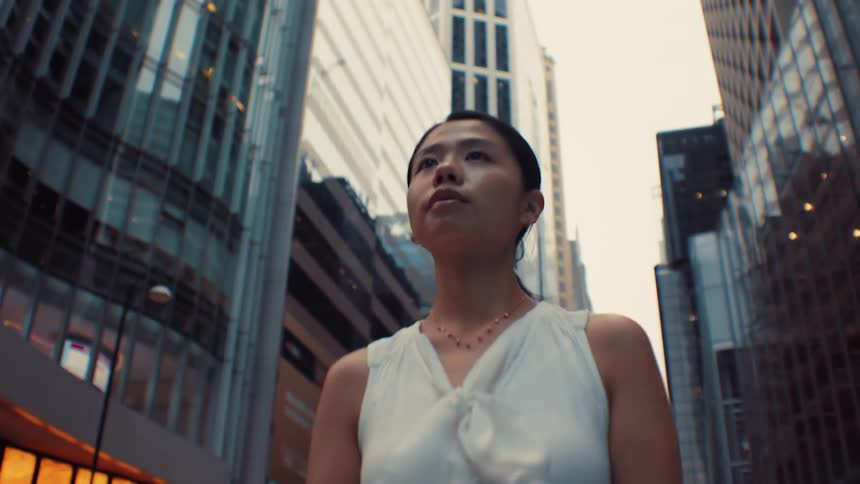 A woman in a white sleeveless blouse walks confidently through a canyon of tall glass skyscrapers in a modern city