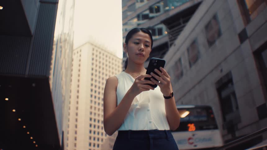 A woman wearing a white sleeveless top and dark pants walks through a city and smiles while looking at her phone with tall buildings and a moving bus in the background