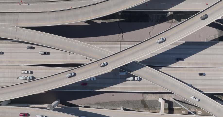 A top view shows many cars and trucks driving along intersecting concrete highway ramps and overpasses on a bright sunny day