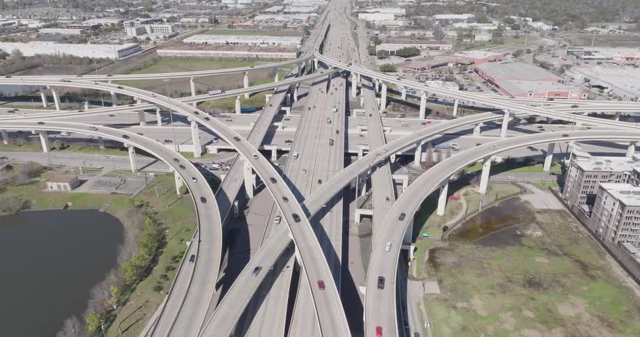 A view from above shows many cars and trucks driving along intersecting concrete highway ramps and overpasses on a bright sunny day
