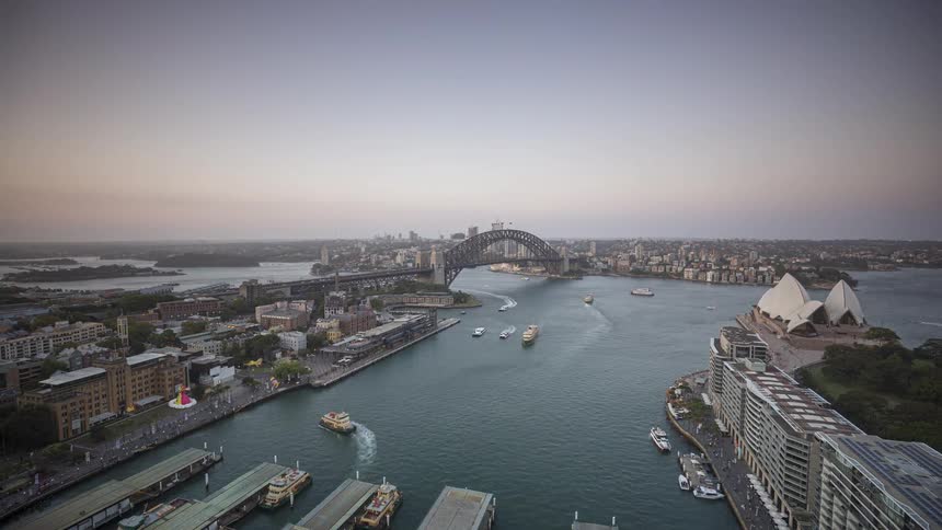 A high angle time lapse view shows the sydney opera house and the sydney harbour bridge as daylight fades and city lights begin to glow under a deepening purple sky while many boats move across the water