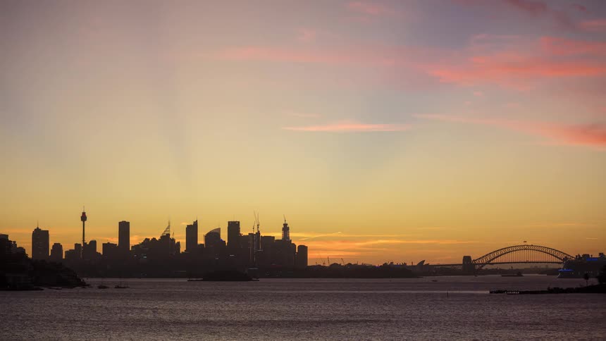Sun sets behind buildings of a metropolitan city on the coast as boats sail on the water with traffic on a bridge in the distance