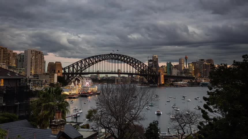 Sailboats sit in a harbor while cars drive across a steel bridge in a metropolis