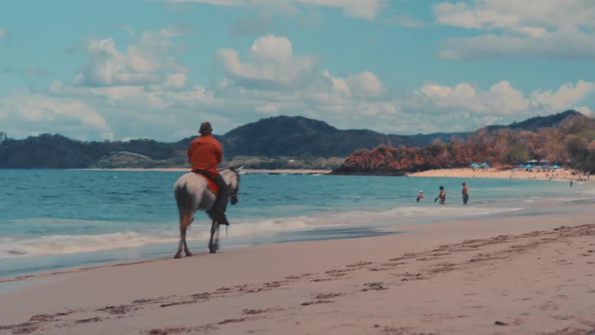 A man wearing an orange shirt and a hat rides a white horse along the shore of a tropical beach while people swim in the distance and clouds hang over the mountains