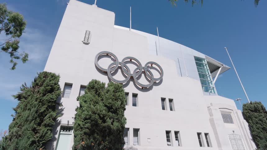 The camera tilts upward to reveal a large silver olympic rings logo mounted in Los Angeles on the side of a modern white buildinga white car drives past in the foreground as the shot captures the architectural details under a clear blue sky