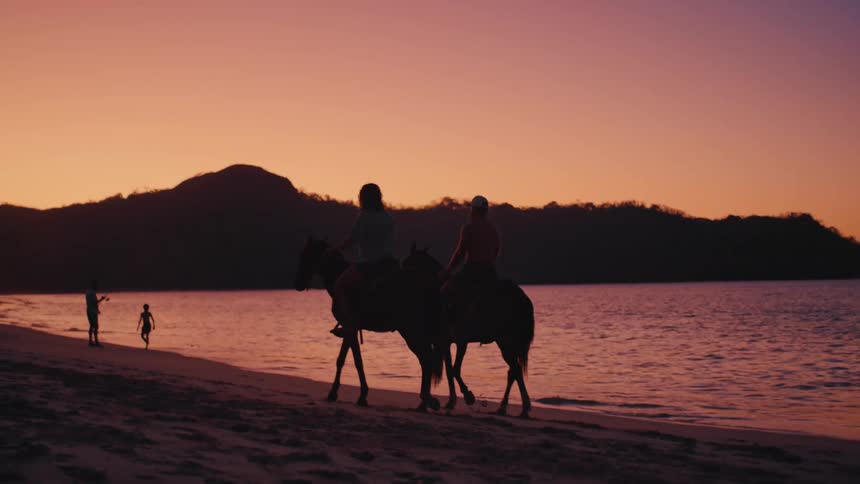 Two people ride horses along a beach as the sun sets casting their figures in dark silhouette against a vibrant orange and purple skyin the background the calm ocean waves lap against the shore while hills rise up behind the distant coastline