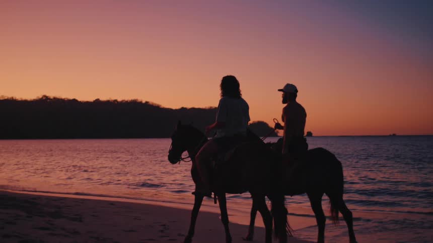 A man and a woman ride horses along a sandy beach at sunset as the orange and purple sky reflects on the ocean water while dark hills sit in the distance