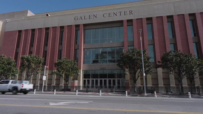 The exterior of the USC Galen Center Olympic Venue featuring its prominent brick facade and glass entrance a white pickup truck drives across the frame in front of the building while the camera remains stationary