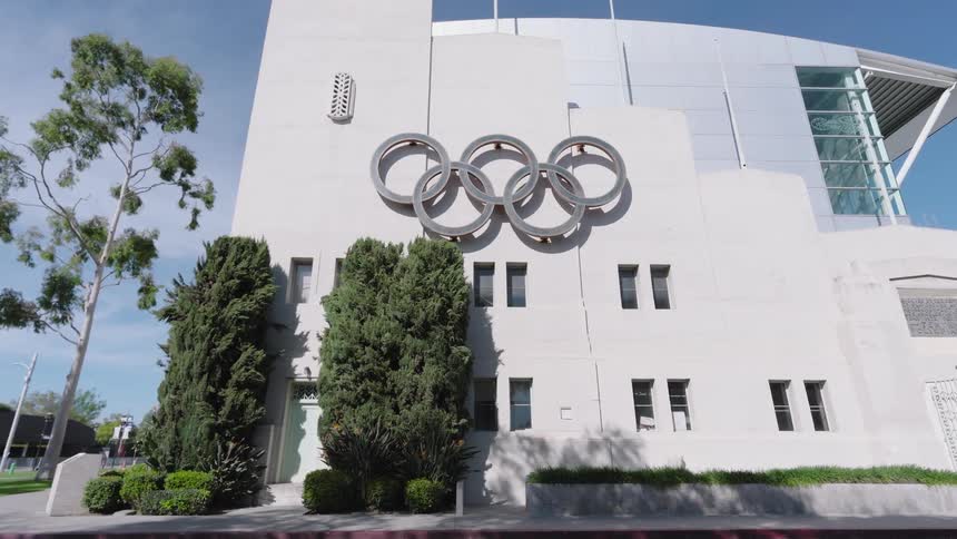 A large silver olympic rings emblem in Los Angeles is mounted on the white facade of a stadium building under a clear blue sky with green trees and parked cars in the foreground during the day