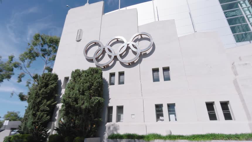An exterior building wall in Los Angeles with the olympic games logo displayed on the side surrounded by trees on a sunny day